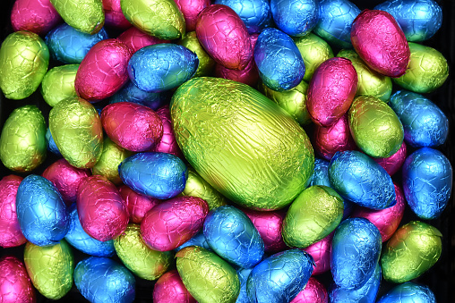 Pile or group of multi colored and different sizes of colourful foil wrapped chocolate easter eggs in pink, blue, yellow and lime green with a large green egg in the middle.