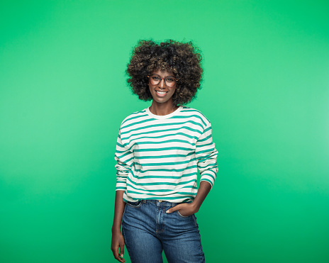 Portrait of funky afro american young woman wearing striped blouse, standing with hands in pockets and smiling at camera. Studio portrait on green background. Easter concept.