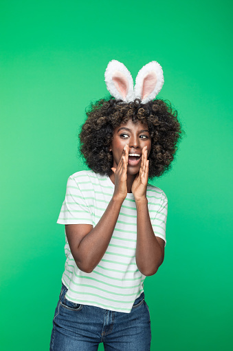 Easter concept. Portrait of excited afro american young woman wearing rabbit ears headband, shouting and raising hands. Studio portrait on green background.
