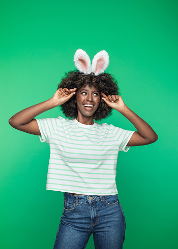 Easter concept. Portrait of excited afro american young woman wearing rabbit ears headband, laughing and raising hands. Studio portrait on green background.