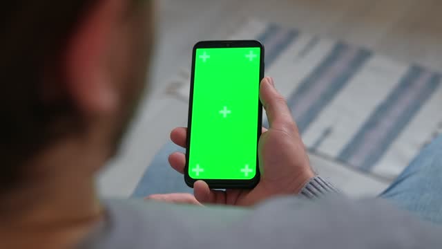 Young man sitting at home holding smartphone green mock-up screen in hand. Male person using chroma key mobile phone. Vertical mode. Touching, swiping display, tapping, surfing Internet social media