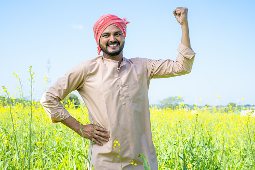 Happy famer showing stroing gesture while standing at farmland by looking at camera - conept of confident, agro business and positive emotion.