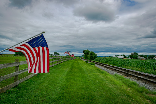 On a Cloudy Day American Flags are Waving in the Wind, on a Fence near a Single Railroad Track