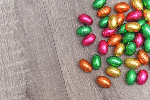 Foil wrapped multi coloured easter eggs in pink, green, orange and yelow in a pile or group, against a grey white wooden background.