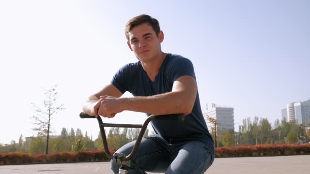 A young man sits on a BMX Bicycle in a Park in Sunny weather.