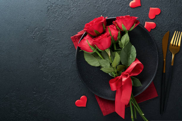 istock Valentines day romantic table setting. Empty closeup of red roses, wine, candles, dinner black plate, knife, fork and decorative silk hearts on black background. Holiday concept. Copy space. Top view 1461096150