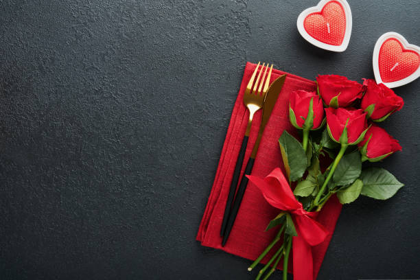 istock Valentines day romantic table setting. Empty closeup of red roses, wine, candles, dinner black plate, knife, fork and decorative silk hearts on black background. Holiday concept. Copy space. Top view 1461096268