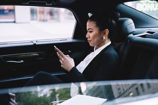 Confident adult Asian executive woman in formal clothes concentrating on screen and interacting with smartphone while sitting in luxurious automobile in city