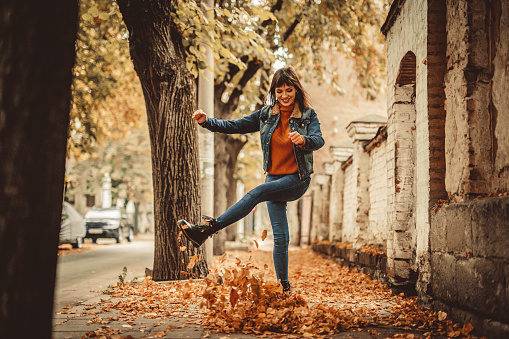 Beautiful young woman kicking a heap of leaves on the street in autumn.