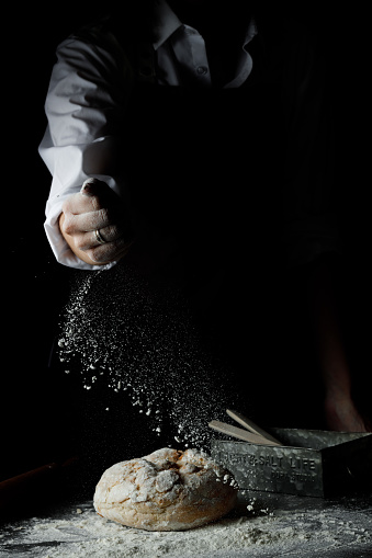 Unidentified woman hand sprinkle, pouring flour to fresh bread on black background. Powdery flour flying into air. Cooking process. Copy space for text