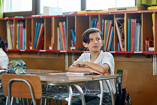 Waist-up view of 10 year old boy wearing uniform, sitting at desk, and looking away at off-camera teacher with expression of uncertainty.