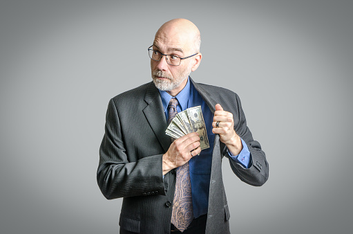Businessman or politician putting money in the inside pocket of his jacket and looking suspiciously sideways on a gray background.