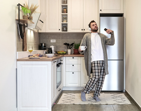 Adult bearded man with headphones holding coffee cup and mobile phone in his hands dancing and enjoying music with eyes closed in domestic kitchen in morning.