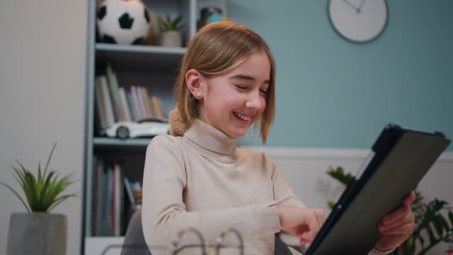 Smart preteen smiling schoolgirl doing her homework with digital tablet at home. Child using gadgets to study. Education and online learning for kids.