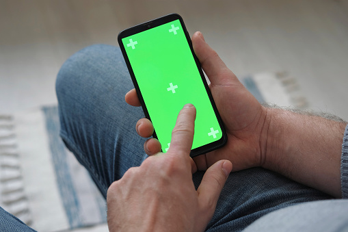 Young man sitting at home holding smartphone green mock-up screen in hand. Male person using chroma key mobile phone. Vertical mode. Touching, swiping display, tapping, surfing Internet social media. Caucasian Man finger clicking, tapping and swiping on center of chromakey green screen. Handsome guy using phone mockup. Close up. Footage Pack. Using for a smartphone, tablet pc or a touch screen devices. Gestures. cellphone with tracking markers. watching content