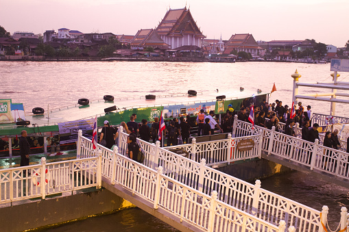 Passenger craft and boat at Yodpiman 2 pier in Bangkok  at sunset of after rain. Thai people are in scene. They are dressed in black at day of funeral of King Rama IX.  Promenade is seated near Buddhayodfa Chulalok Maharat Bridge