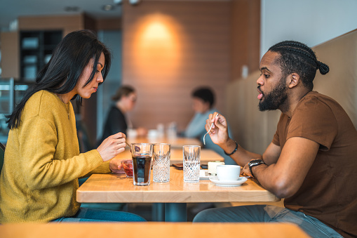 Waist up shot of mid adult African man and South American woman sitting at a lunch restaurant, talking while eating. Side view.