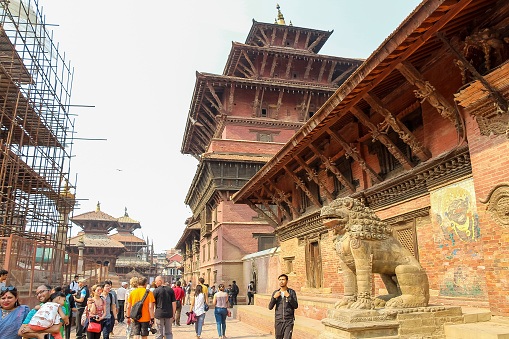 Nepal, Lalitpur - May 10, 2019: People walks by stone guardian lion statue standing by entrance of hindu Taleju temple on Patan Durbar Square in a sunny day. Religious architecture theme.