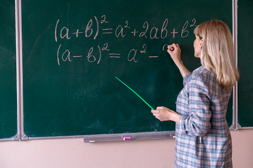 A young Caucasian female math teacher stands at the blackboard and explains the solution of a mathematical problem