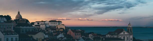 A panoramic view of an antique and typical Lisbon city with modest housing, churches, and the magnificent river with a stunning sunset in the background