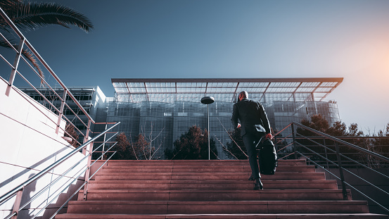 A sophisticated African man executive was captured from behind walking up a large staircase toward an architecturally beautiful and innovative glass made office building