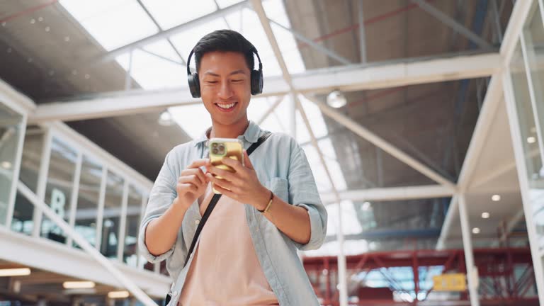 Phone, music headphones and Asian man walking in mall streaming podcast or radio. Technology, travel and happy male listening to song, audio sound or playlist on mobile smartphone in urban building.