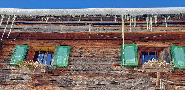 Two small window on wooden mountain cottage in winter. Icicles hanging from the roof against blue sky. f