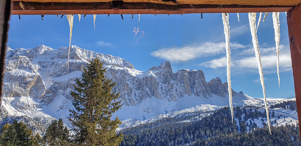 View through the window at mountain peaks in Col Rodella Ski arena, Dolomites. Long icicles hanging from the roof on beautiful sunny day.