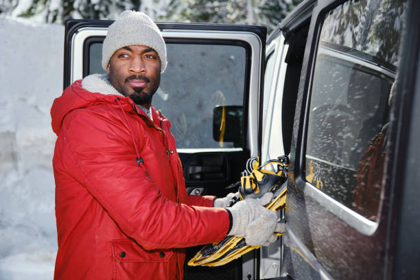 Adult man in winter clothing with knitted hat puts something into white car trunk in snowy day. Winter season concept.