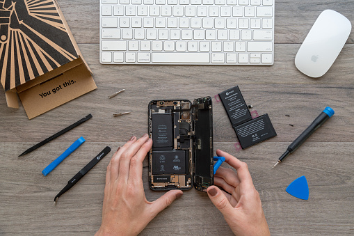 A woman at her desk performs a battery replacement on an iPhone X using an iFixit self repair kit. Kits like these include tools like precision screwdrivers with bits to remove proprietary screws, opening picks, pointed tweezers, suction cups, and spudgers to empower regular users to repair their own devices.