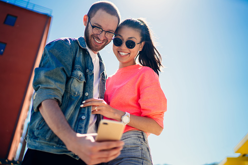 Below view of happy bloggers clicking smartphone pictures during leisure connecting to 4g internet for sharing influence content to social networks, joyful teenagers smiling at front cellular camera