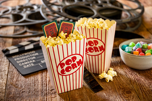 This is a conceptual photo relating to going out to the movies or watching a movie at home. There are two old retro movie reels on a wood background with two bags of popcorn and hand made cinema tickets.
