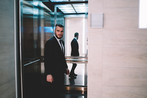 Stylish adult serious confident man in elegant suit standing in opened elevator of commercial building and looking away over shoulder