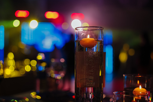 Banquet table decorated with burning candles in glass vases in restaurant hall. In the background party with silhouettes of people dancing on the dance floor with disco lights glowing searchlight.