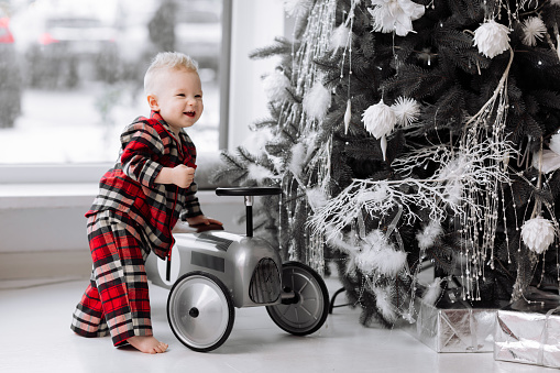 adorable smiling baby boy in costume in red checked plays with toy retro grey car by festive decoration Christmas tree and big window. Happy childhood. Happy family Xmas and New Year holiday vacation
