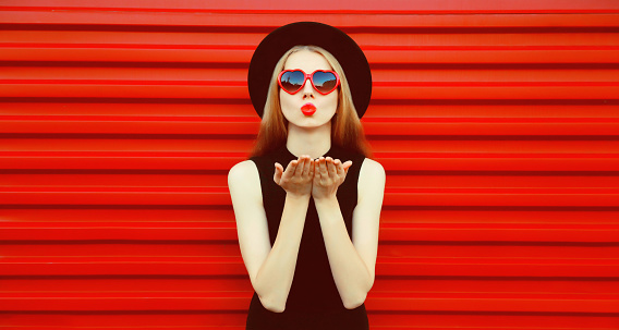 Portrait of beautiful young woman blowing her lips with lipstick sending sweet air kiss wearing red heart shaped sunglasses, black round hat on background