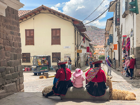Cusco, Peru- January 2023: Peruvian women with traditional clothing and street with people,  souvenir shops and view of mountain with houses.