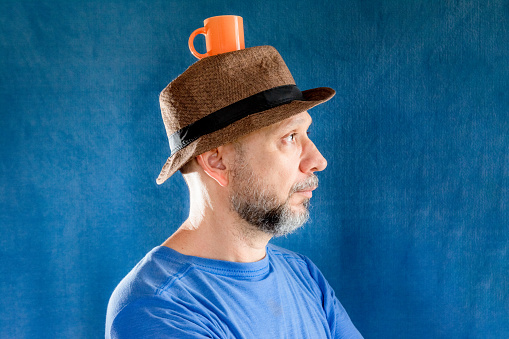 Studio portrait of a bearded man, wearing a hat, with an orange cup on top of the hat. Isolated on blue background.