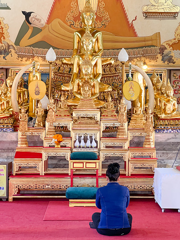 Back view of unrecognizable female in casual clothes sitting near golden Buddha statues and worshipping during visit to oriental temple