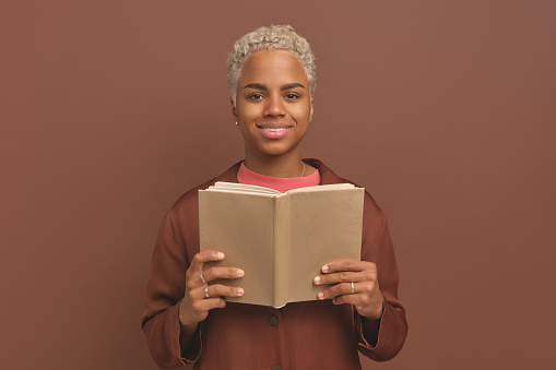 Young optimistic African American woman gen z holding open book in front of chest and looking at camera rejoicing at opportunity to re read novel of favorite author stands on brown studio background