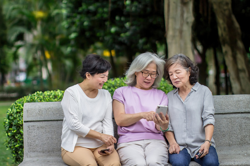 A heartwarming scene of three senior women sitting on a bench, engrossed in the screen of a smartphone. With smiles on their faces, they share a moment of modern connection and camaraderie, proving that age is no barrier to embracing technology and forming lasting friendships.