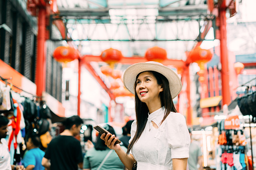 Female tourist holding smartphone looking around Petaling streets, Kuala lumpur's Chinatown area during the day.