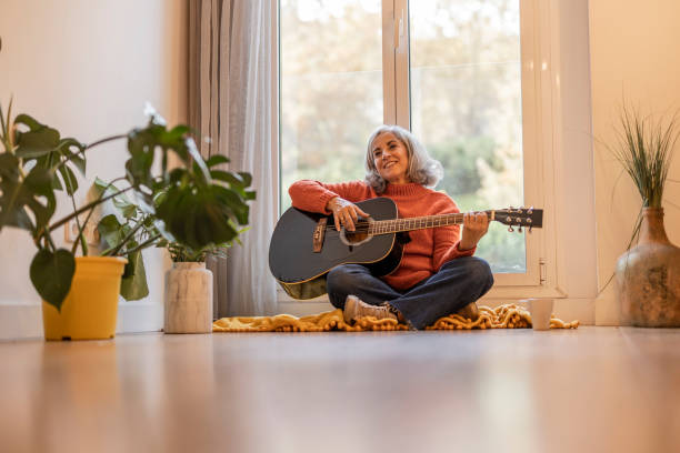 femme souriante jouant de la guitare - femme mûre aux cheveux gris prenant des cours de guitare en ligne à la maison - - musician music women guitar photos et images de collection