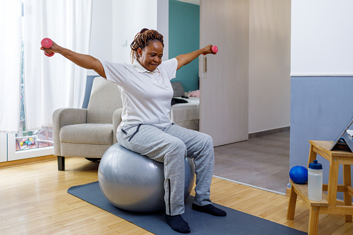 African woman doing workout with dumbbells at home while watching online tutorial.