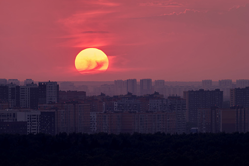 Big red sun over the roofs of city buildings, evening sunset. Cloudy sky and the sun setting behind the houses