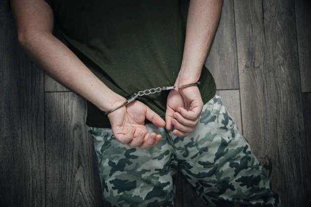 istock A military soldier in handcuffs, hands behind his back, on a dark background. Concept: war criminal, prisoner of war, tribunal. capture of a prisoner in the war. 1461272476