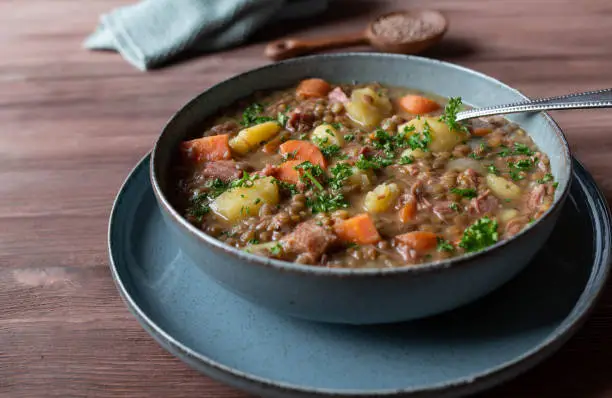 Delicious and healthy homeade lentil soup or lentil stew. Traditional german recipe. Cooked with brown lentils, potatoes, root vegetables such as carrots, onions and  celery. Topped with parsley. Served isolated on wooden table. Closeup and front view