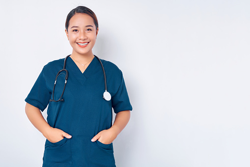 Smiling young Asian woman nurse wearing blue uniform with stethoscope holding hands in pockets isolated on white background. Healthcare medicine concept