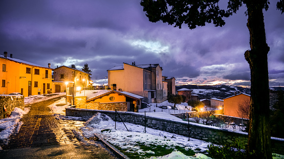 A winter atmosphere at twilight in the snowy alleys of the medieval village of Gualdo Tadino in Umbria, central Italy. The Umbria region, considered the green lung of Italy for its wooded mountains, is characterized by a perfect integration between nature and the presence of man, in a context of environmental sustainability and healthy life. In addition to its immense artistic and historical heritage, Umbria is famous for its food and wine production and for the high quality of the olive oil produced in these lands. Image in 16:9 ratio and high definition quality.