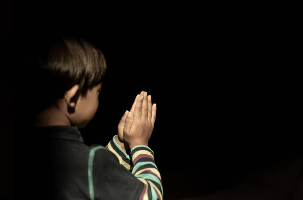 Child praying with joined hands isolated on black background, turned from behind unrecognizable
